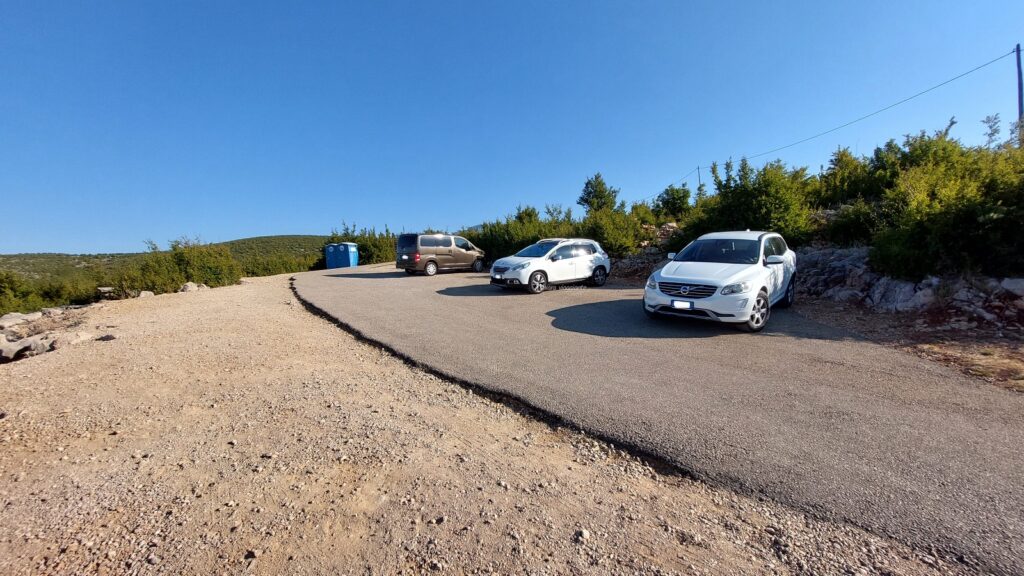 parcheggio asfaltato vicino all’imbocco del sentiero per raggiungere Kudin Most, con auto e vegetazione circostante.