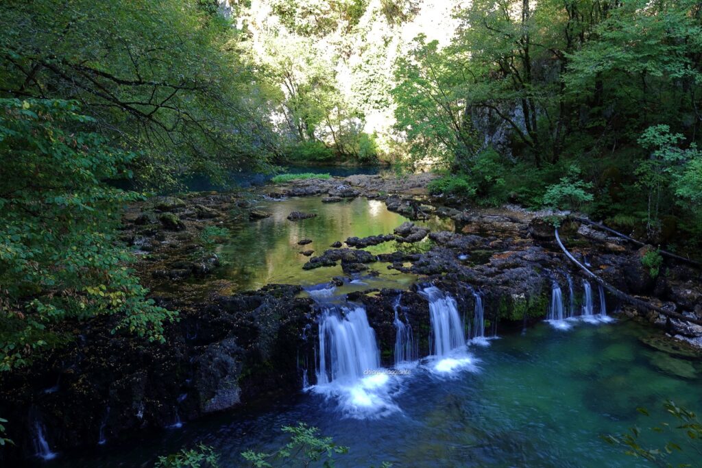 Piccola cascata alla sorgente del fiume Una, con acqua limpida che scorre tra rocce e vegetazione del bosco.