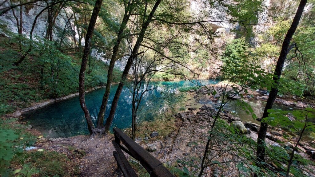 Lago turchese della sorgente dell’Una visto dalla fine del sentiero, con acqua limpida circondata da rocce e vegetazione.